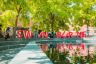 Students sitting near large red Swinburne letters beside a reflective pond under leafy trees.